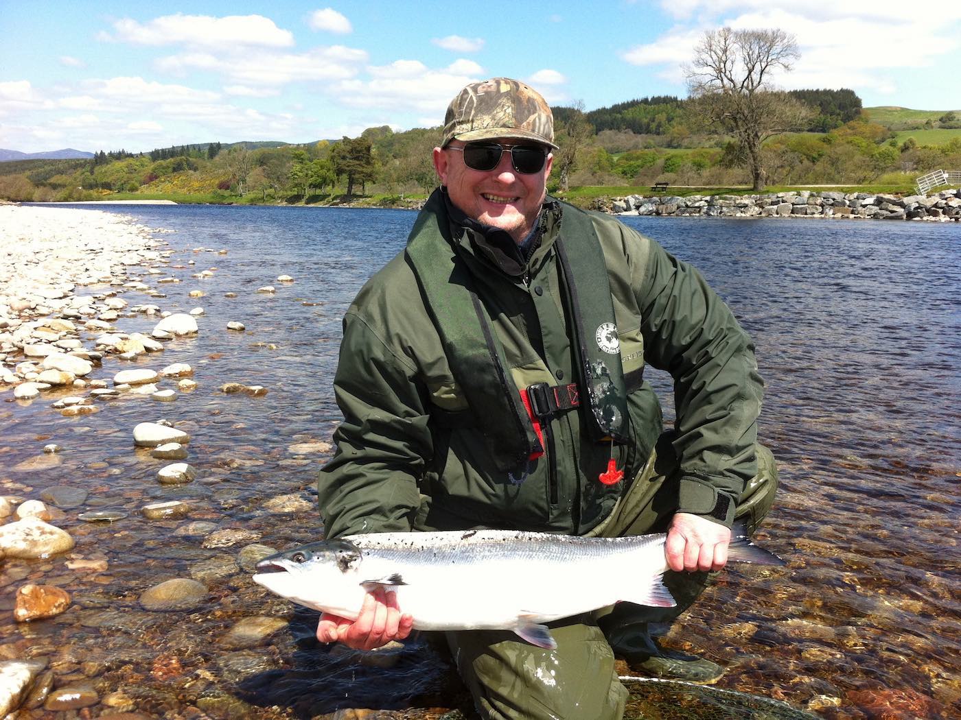Look at the beautiful silver flanks of the June caught river Tay fresh run salmon which was hooked and landed at the Ash Tree Pool on The Lower Kinnaird Beat of The River Tay on this bright light early June fishing day. Note the salmon fishing trip guest is wearing eye protection and a life jacket as safety always comes first on the salmon river.