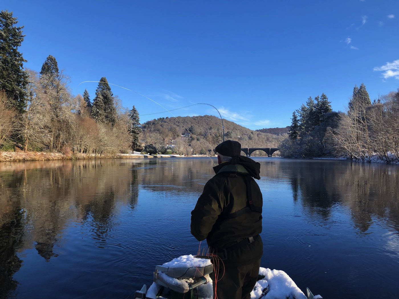 This River Tay salmon fisher is Spey casting from the traditional Tay salmon fishing boat during late January on the Tay at Dunkeld with his 15ft double handed salmon fly rod. If you look in the distance you'll see the 200 year old Telford Bridge which is one of the Tay's most historic & beautiful sandstone bridges.