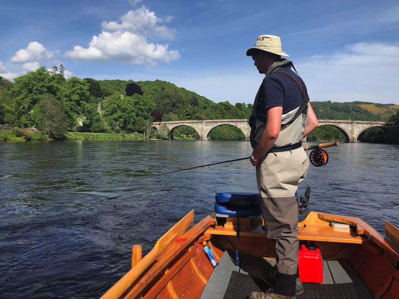 Here's a salmon fly fisher out in a traditional River Tay coble boat covering the Cathedral Stream salmon pool with a fly rod. This is a great area for a resting salmon to be located that's navigated up through the fast water below the bridge. The Telford Bridge you can see in the shot is over 200 years old and joins the village of Birnam to the town of Dunkeld.